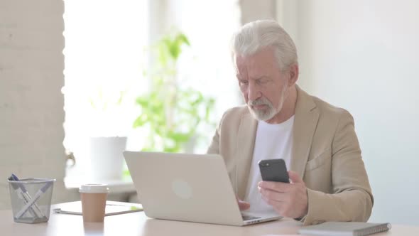 Old Man Browsing Internet on Smartphone While Using Laptop in Office