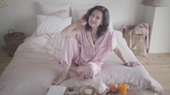 Portrait of Happy Caucasian Woman in Nightwear Dreaming As Sitting on Bed at Home. Smiling Brunette