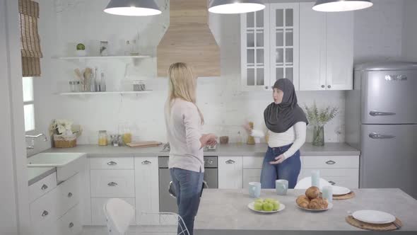 Muslim Woman in Hijab Arguing with Blond Caucasian Friend at Home. Two Women Quarrelling Indoors