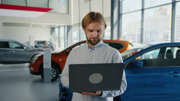 Bearded Attractive Male Manager with Laptops on the Background of a Car Dealership