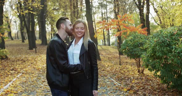 Happy Couple Stands in Embraces and Man Whispers in Girl's Ear in Park