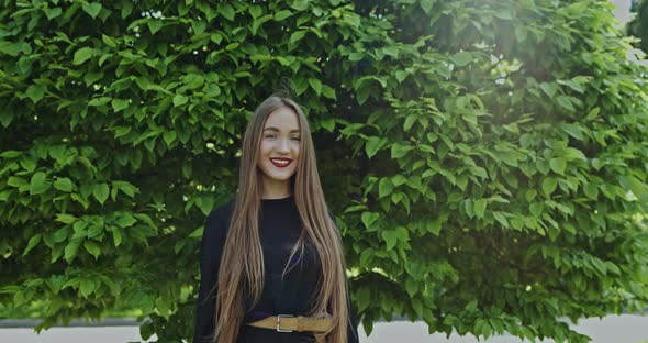 Young Smiling Woman in Black Dress and with Long Gorgeous Hair Standing Against Lush Green Tree in