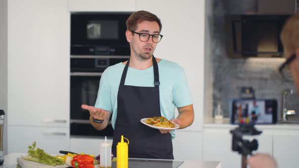 Young Man Holding Plate with Pasta and Telling How To Cook Vegetarian Dish on Camera in Kitchen