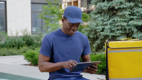 Smiling Afroamerican Man Courier Food Delivery Sitting on Bench Uses Tablet