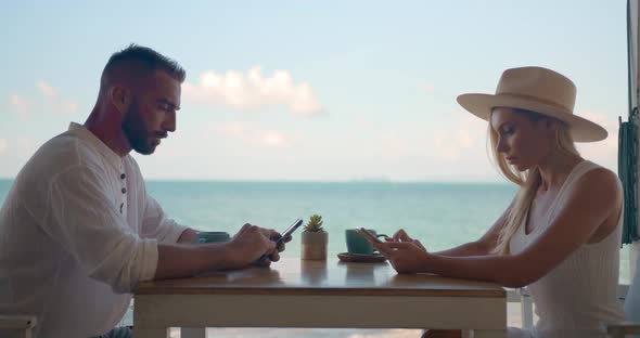 Young Man and Woman Sitting in Cafe By the Sea Both Using Their Smartphones to Check Morning News