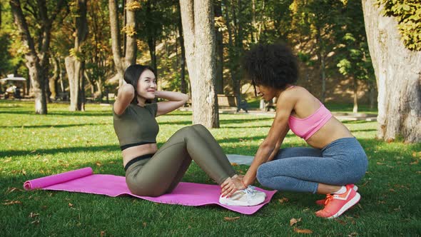 Smiling African American Woman Supporting Her Asian Friend Lying on Mat for Doing Crunches at Park