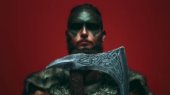 A Viking in war paint leans his beard on an axe, opens his eyes and looks aggressively at the camera