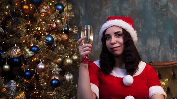 Young Woman in Santa Claus Costume with Glass of Champagne Sitting on Chair Near Christmas Tree