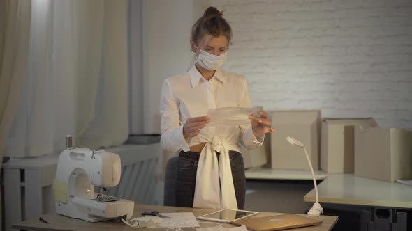 Portrait of Caucasian Seamstress Standing in Atelier and Looking at Covid-19 Face Mask Packed in