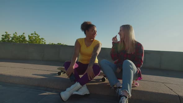 Pretty Diverse Female Friends Sitting on Skateboards and Chatting Outdoors