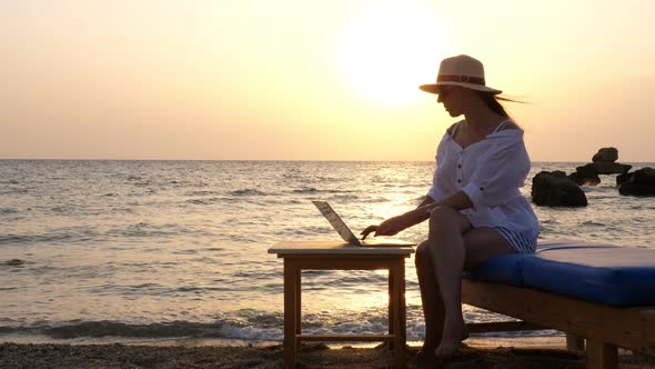 Young Woman in Sunglasses and Sun Hat, Uses Laptop, Sitting on the Beach By the Sea, at Sunset or