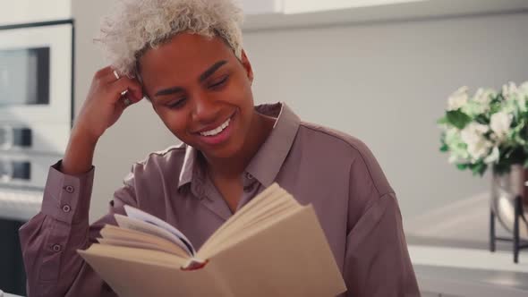 Happy African American Woman Sitting Reading Interesting Book in Kitchen