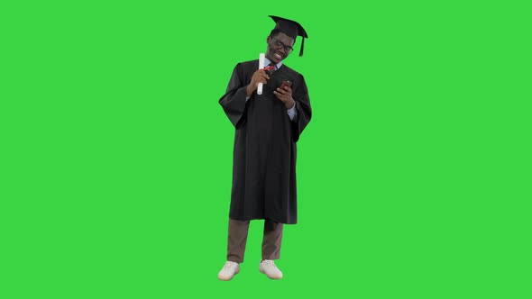 Smiling African American Male Student in Graduation Robe with Diploma Texting on the Phone