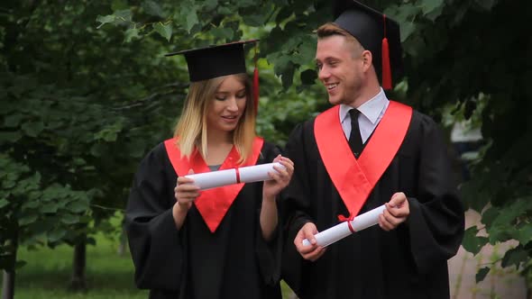 Woman and Man in Academic Dresses Holding Diplomas Talking and Walking in Park