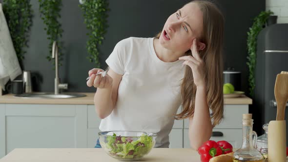 Unhappy Woman Eating Vegetable Salad at Table in Kitchen