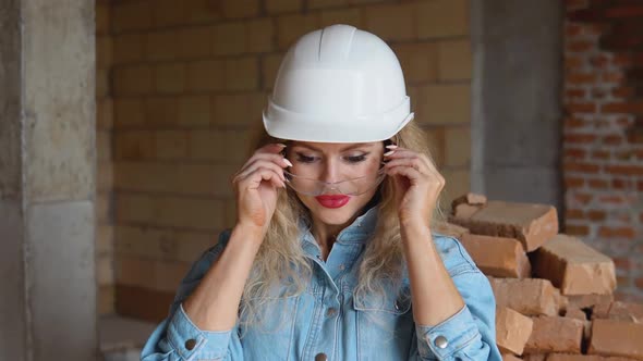 Young Woman in a Work Helmet and Denim Clothes Wears Glasses at the Construction Site