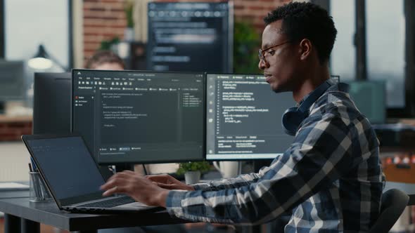 Portrait of African American Developer Using Laptop to Write Code Sitting at Desk with Multiple