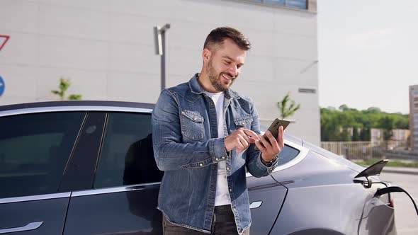 Bearded Man Using His Phone while Charging His Luxurious Electromobile in Charging Station