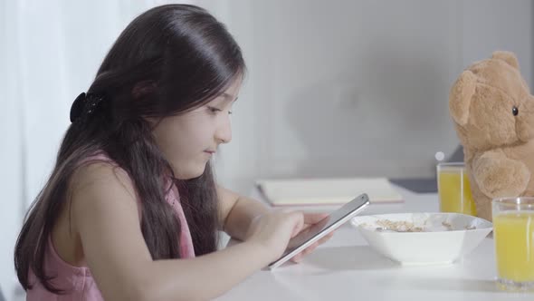 Side View of Concentrated Little Middle Eastern Girl Using Social Media During Breakfast. Cute Child