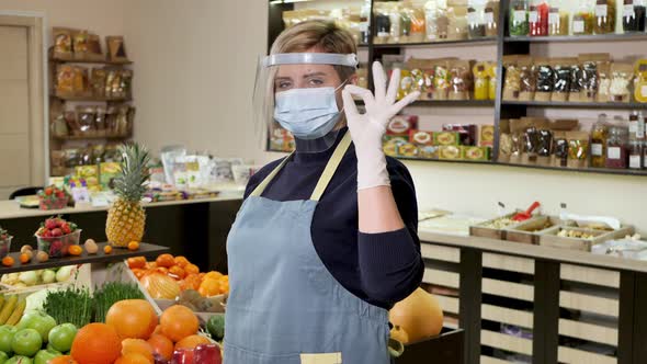 Young Female Health Food Store Clerk Wearing Gloves and a Protective Mask Looks at the Camera and