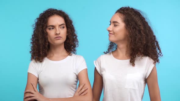 One Dissapointed Female Curly Caucasian Twin Standing on Blue Background While Another Smiling