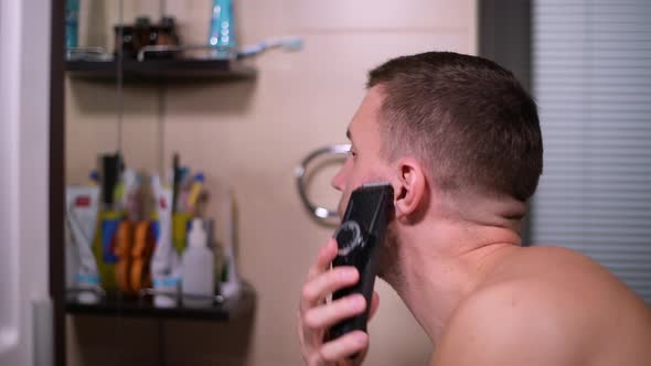 a man cutting his beard with a trimmer in front of a mirror