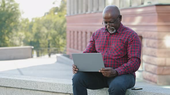 African American Mature Man In Shirt Sitting Outdoors Using Laptop Communicating on Social Networks