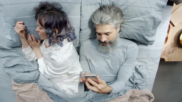 Mature Man Husband Using Smartphone Texting While Young Woman Sleeping in Bed