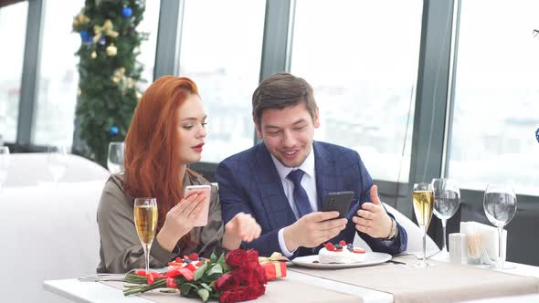 Young European Couple on a Date in Restaurant. Red-haired Girl Shows a Guy Pictures on His