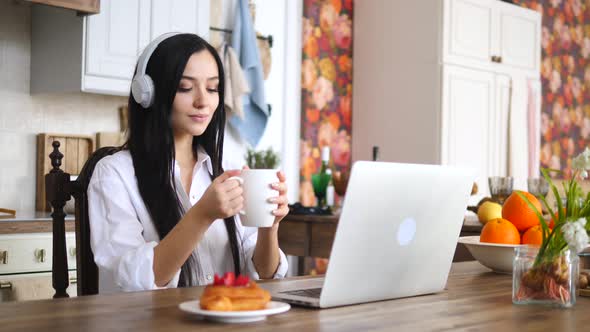 Young Smiling Woman Using Laptop Computer And Holding Cup Of Tea Or Coffee On Kitchen.