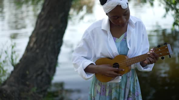 Concentrated Talented African American Woman Playing Ukulele Standing on River Bank in Spring Summer