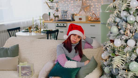 Woman in Santa Hat Sitting on Sofa Near Christmas Tree and Scratching Head