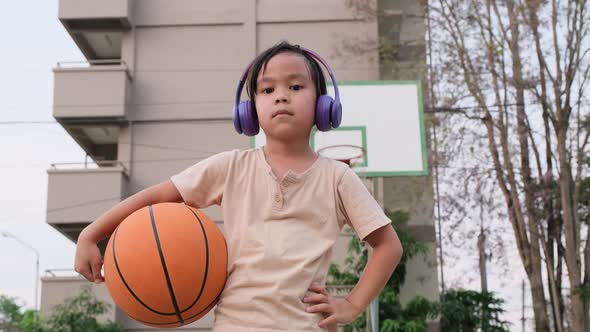 Cute little girl wearing headphones poses with basketball at outdoor basketball court. alt
