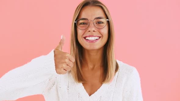Young Woman in Glasses and White Jumper is Showing Thumb Up Smiling While Posing on Pink Background
