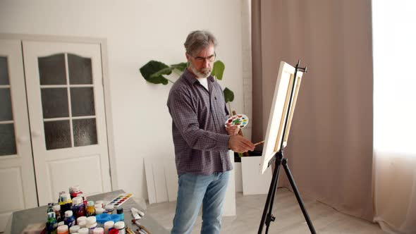 Man Holds Palette in Hand and Draws with Brush on Canvas Standing in Living Room There are Many Cans