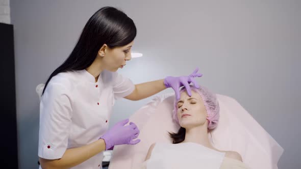 A beautiful cosmetologist advises a client who lays on a chair and examines her facial wrinkles