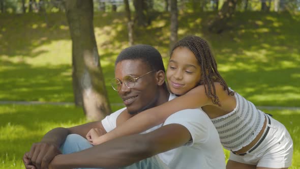 Side View of Cute Girl Hugging Man in Sunny Summer Park. Portrait of Happy African American Sister