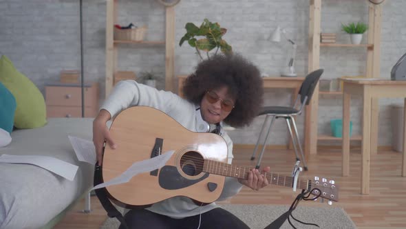 Cheerful African American Woman with an Afro Hairstyle Plays Guitar Sitting on the Floor