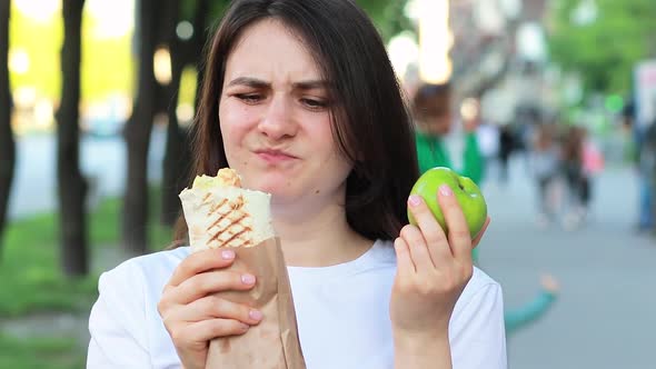 Young Beautiful Brunette Woman on a Diet Chooses Food  What to Eat an Apple or Fast Food Shawarma