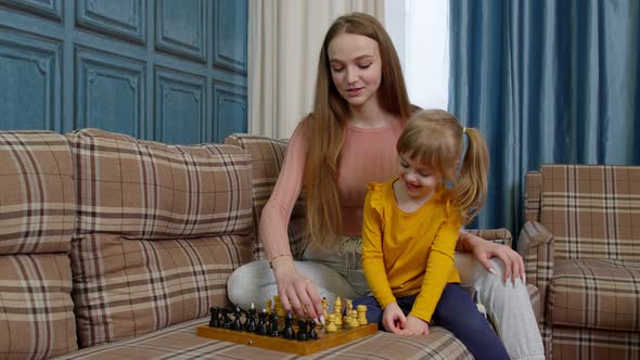 Happy Young Mother Woman Teaching Small Child Daughter Playing Chess on Wooden Board at Home Sofa