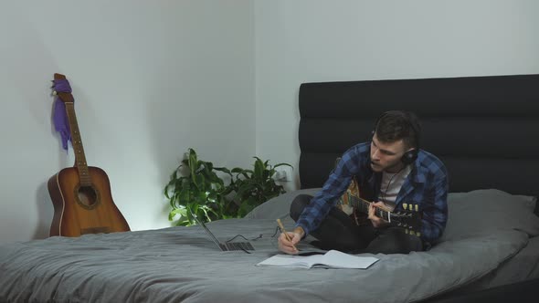 Young man in headphones creating song, taking notes and playing on electric guitar. Music concept