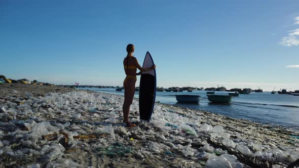 Caucasian young athletic female surfer stand in polluted beach with plastic waste trash garbage, Mot