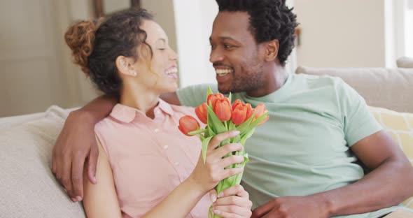 Happy biracial couple sitting on sofa with bouquet of tulips