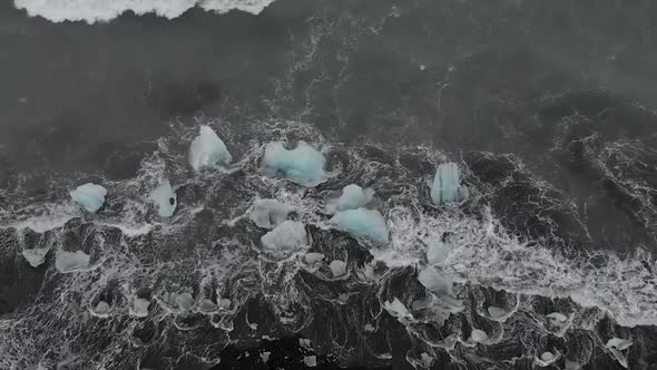 Slow motion panning drone shot of waves crashing into black sand beach icebergs.