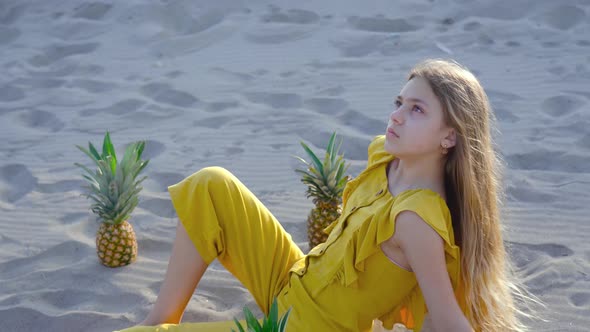 Young Blonde Girl Sitting on the Sand Among Pineapples