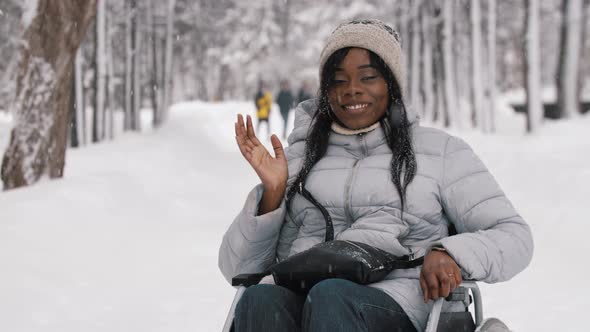 Young Black Woman Sitting in a Wheelchair in Winter Forest and Waving Her Hand