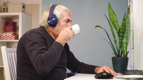 Senior Man in Headphones Seated at Table Looking at Laptop