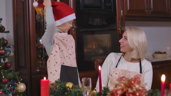 Positive Little Son in Apron Putting on Christmas Hat Cheerful Mother Showing Thumbs Up and Smiling