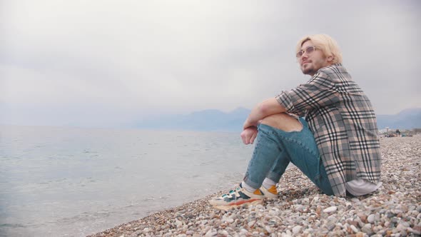 Smiling Young Man in a Plaid Shirt Sits on the Shore