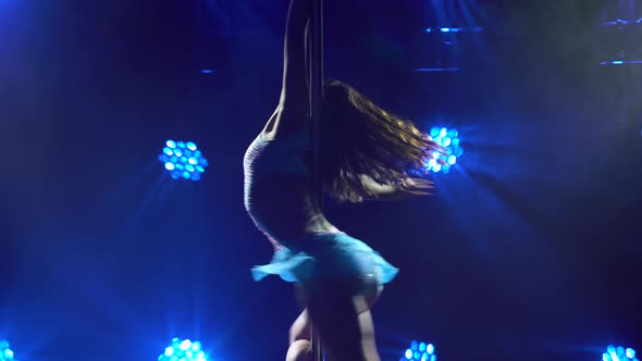 Athletic Flexible Woman Rotates on a Pole in a Dark Studio with Blue Neon Lights